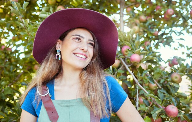 Trendy farm worker wearing a hat daydreaming on a fruit farm during harvest season Young smiling farmer in front of a apple tree on a sunny day The agricultural industry growing fresh produce