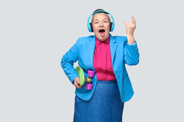 Trendy funny grandmother in casual style with blue headphones holding green skateboard listen music showing rock sign tougue out, looking at camera. Indoor, studio shot, isolated on gray background