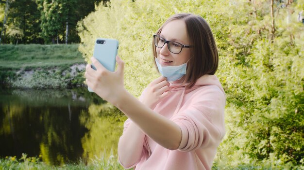 保護用の医療用マスクと眼鏡をかけたトレンディな女の子がスマートフォンで自分撮りをします