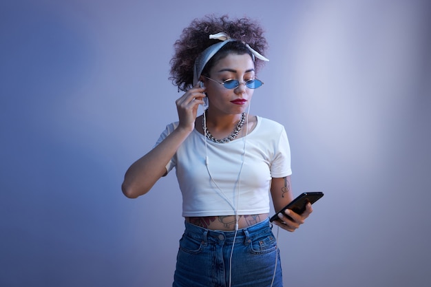 Trendy girl with afro curls uses a smartphone and earphones on a blue studio background with copy space, online conversation, listens to music