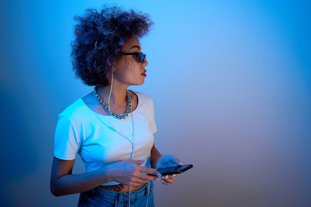 Trendy girl with afro curls uses a smartphone and headphones on a studio background with copy space