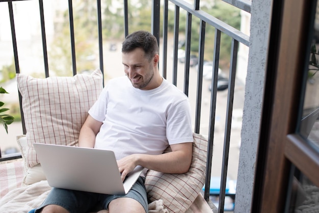 Trendy happy caucasian man sitting at balcony while working remotely on laptop