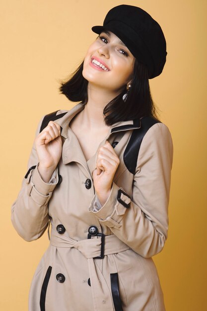 Модная счастливая женщина в элегантном пальто и черном берете, улыбаясь в камеру на коричневом