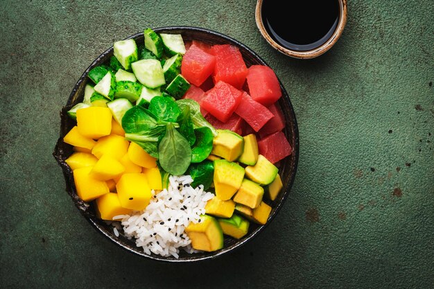 Trendy hawaiiaanse poke bowl met tonijn avocado mango komkommer veldsla en witte rijst sojasaus en sesam dressing oude groene tafel achtergrond bovenaanzicht