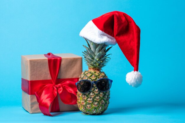 Trendy hipster ananasfruit met zonnebril en een rode kerstmuts naast een geschenkdoos met een rood lint op een blauwe achtergrond