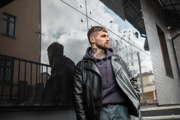 Alla moda hipster uomo hipster con acconciatura e tatuaggi in giacca di pelle alla moda rock nero e felpa con cappuccio cammina vicino a un moderno edificio nero sulla strada