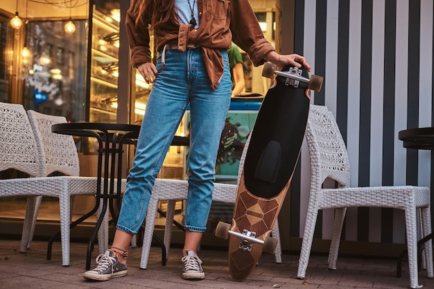 Trendy hipster vrouw poseert voor fotograaf met haar longboard in de buurt van café.