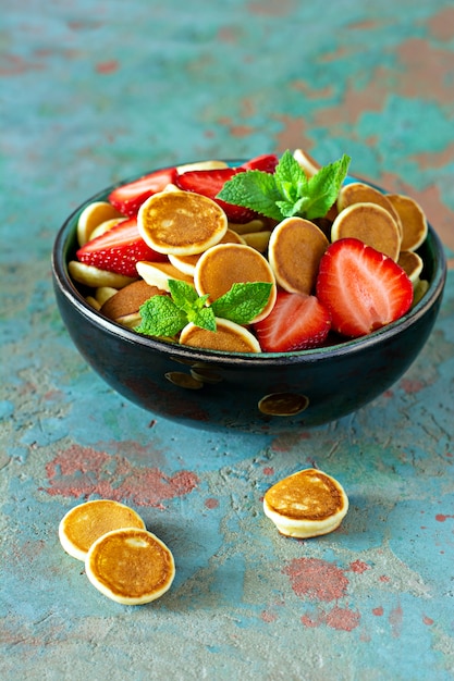 Trendy home breakfast with tiny pancakes (mini pancakes) with strawberries and mint in a bowl (cup) on a blue background.