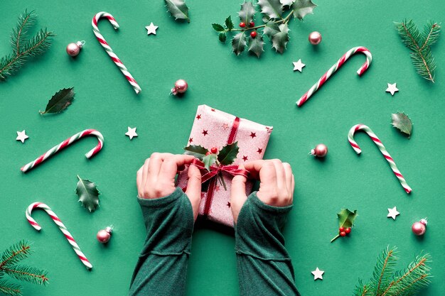 Trendy kerstplateau met snoepriet, hulst- en vuurtakjes, houten sterren en glazen snuisterijen. Vrouwelijke handen binden lint op geschenkdoos verpakt in roze inpakpapier.