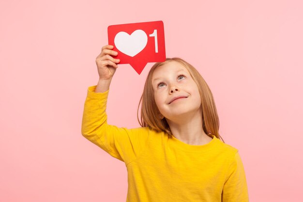 Trendy kind inhoud bloggen portret van schattig voorschoolse meisje kijken naar sociale media hart als pictogram op haar hoofd volger kennisgeving symbool indoor studio shot geïsoleerd op roze achtergrond