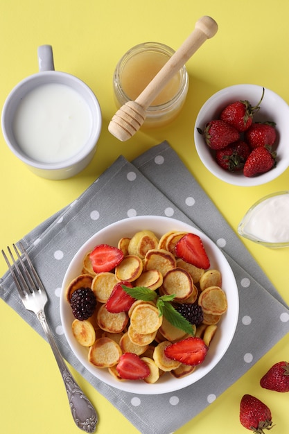 Trendy kleine pannenkoeken voor het ontbijt met aardbeien, bramen en honing in witte kom op gele achtergrond.