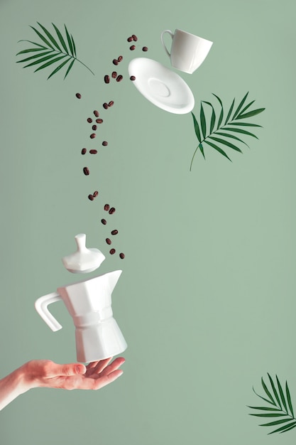 Trendy levitation. Flying coffee beans and espresso cup with saucer. Green mint wall with palm leaves, text space. Ceramic stove coffee maker balancing on index finger of female hand.