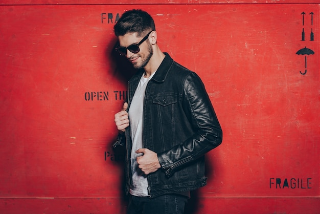 Trendy look. Handsome young man in sunglasses adjusting his jacket and looking away with smile while standing against red background