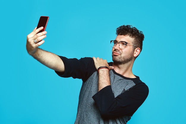 Trendy man using phone and taking selfie on blue backdrop