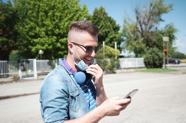 Trendy man with headphones around neck and lowered surgical\
mask using mobile phone outdoor after coronavirus outbreak happy\
man doing video call with smartphone after lockdown new normal\
concept