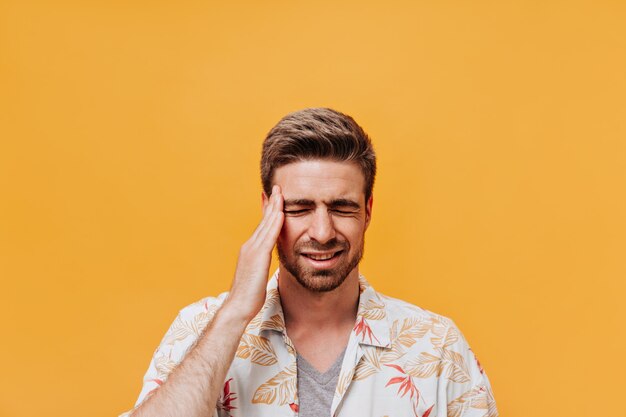 Trendy man with stylish beard in white summer printed shirt and grey t-shirt with headache posing with closed eyes on orange wall