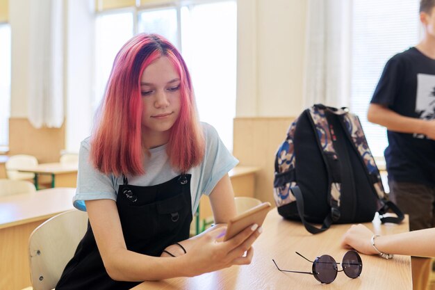 Trendy meisje tiener schoolstudent, in de klas met smartphone in de hand, zittend aan bureau tijdens pauze tussen lessen