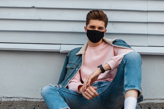 Trendy model young man in stylish denim jacket in torn fashionable blue jeans in fashion black mask sit on stone tile near vintage wall in city. Attractive guy in trendy casual clothes on street.