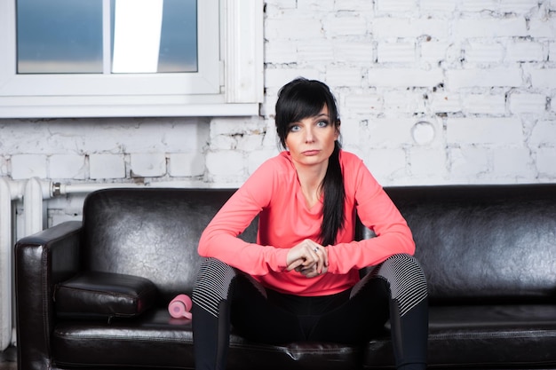 Trendy mooi meisje brunette in lichte sportkleding zit op de bank en kijkt in de camera klaar om te beginnen met trainen Het concept van sportfitness