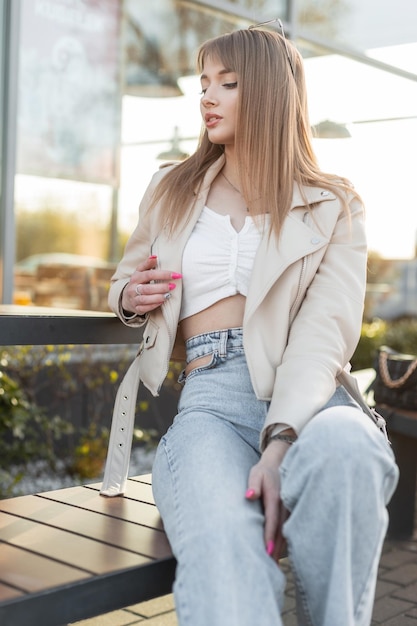 Trendy mooie jonge mode hipster meisje met haar in modieuze stijlvolle rock kleding met een leren jas top en vintage spijkerbroek zit op een houten bankje in de buurt van een café bij zonsondergang
