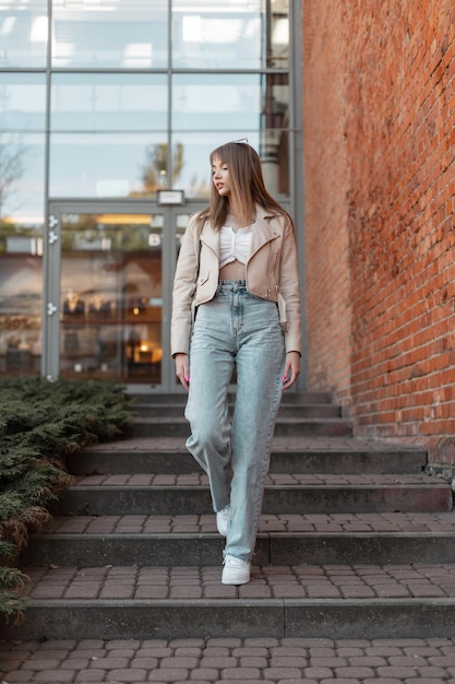 Trendy mooie jonge vrouw in modieuze vrijetijdskleding met een leren jas met jeans met wijde pijpen en witte sneakers loopt in de buurt van een winkelgebouw van rode baksteen