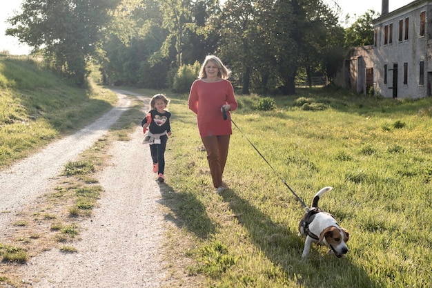 Trendy senior lady and her little toddler girl enjoying a walk in the countryside with their beagle dog Grandmother and granddaughter happy together in the park in sunny day Relationship concept