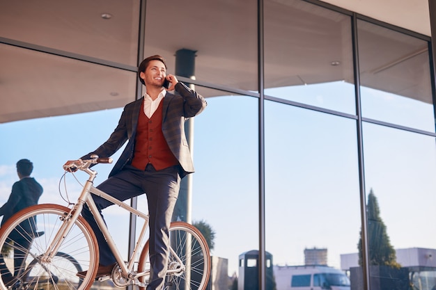 Photo trendy smiling businessman talking on phone sitting on bicycle