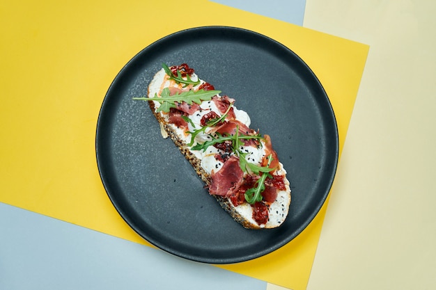 Trendy straatsnack. Lekkere sandwich met jamon, gepocheerde eieren, komkommer en microgreen op zwarte plaat op gekleurde oppervlak kopie ruimte.