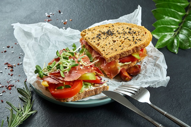 Trendy street snack. Tasty sandwich with jamon, tomatoes, cucumber and microgreen on craft paper on a black surface