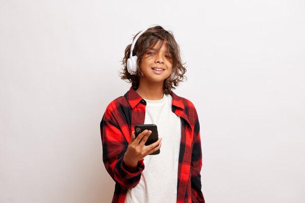 Trendy teen guy listens music through wireless headphones and\
enjoy life stands in front of a white wall