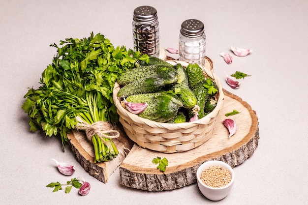 Trendy ugly organic cucumbers in a basket. Fresh vegetables, garlic cloves, mustard, parsley bunch, spices. Wooden stand, modern hard light, dark shadow. Stone concrete background, copy space