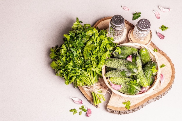 Trendy ugly organic cucumbers in a basket. Fresh vegetables, garlic cloves, mustard, parsley bunch, spices. Wooden stand, modern hard light, dark shadow. Stone concrete background, top view