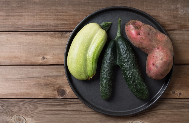 Trendy ugly organic natural potato,cucumber and squash on the black plate, on natural wooden table. Copy space.