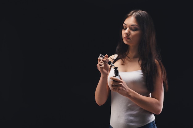 Trendy vaping gadget. Young brunette girl filling vape mod with fruit nicotine free e-liquid at black background, studio shot. Modern device for smokers, copy space