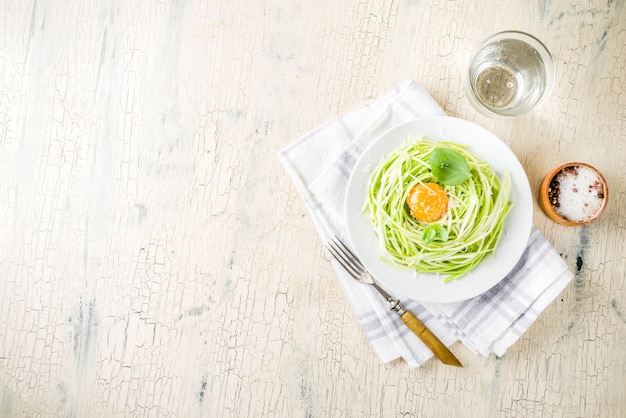 Trendy vegan food recipes, cheese zucchini spaghetti pasta with egg yolk with parmesan, olive oil and basil leaves, light concrete background 