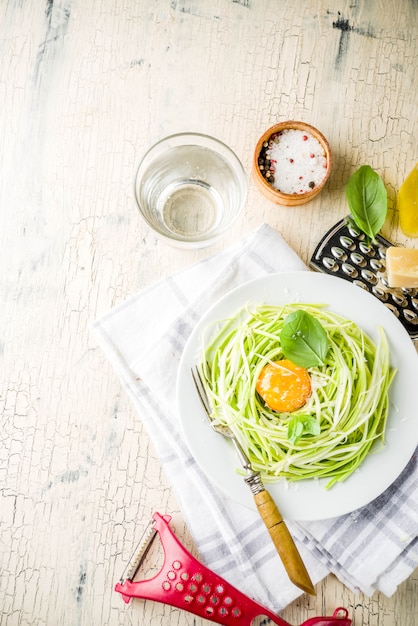 Trendy vegan food recipes, cheese zucchini spaghetti pasta with egg yolk with parmesan, olive oil and basil leaves, light concrete surface 