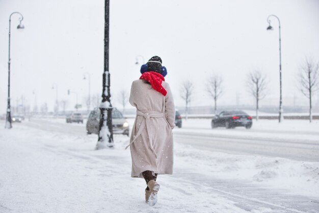 Trendy woman in beige coat, red scarf walking on empty street during snowfall