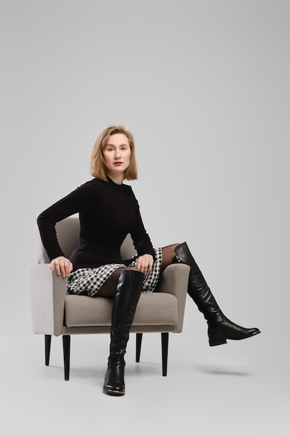 Trendy woman in turtleneck and suspender belt over little skirt sitting in armchair in studio over grey background