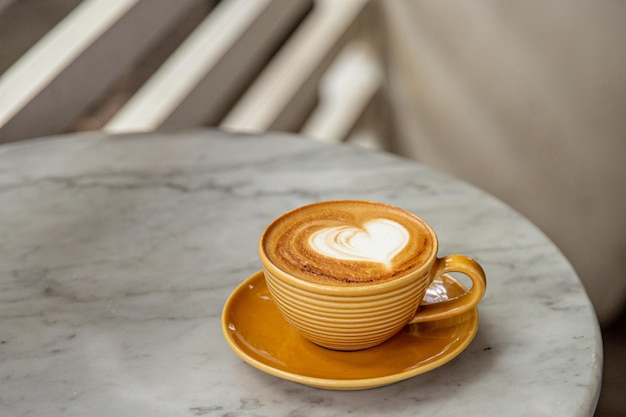 Trendy yellow cup of hot cappuccino on marble table background Heart shape latte art for symbol of love