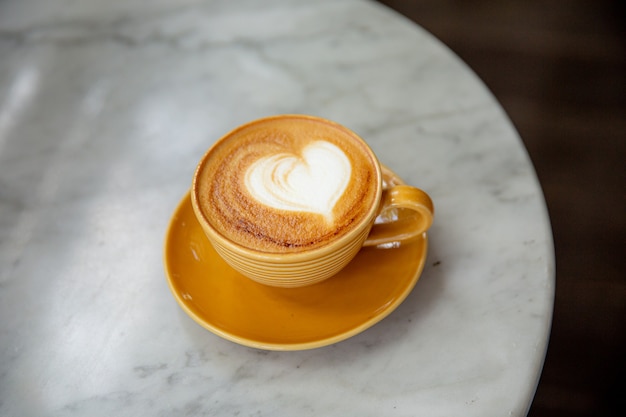 Trendy yellow cup of hot cappuccino on marble table Heart shape latte art for symbol of love