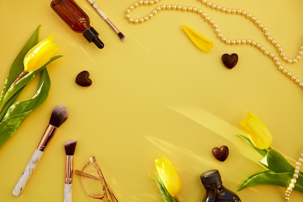 Trendy yellow feminine flat lay makeup brushes glasses serum dropper pearls chain Golden accessories chocolate hearts and yellow tulips Beauty card concept Copy space