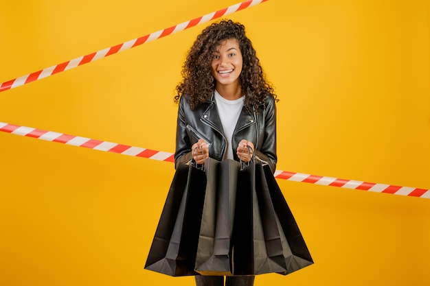 Trendy young black woman wearing jacket with paper shopping bags isolated over yellow with signal tape