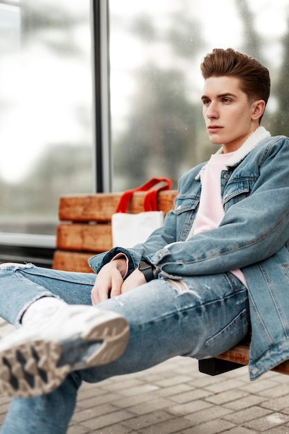 Trendy young man in youth stylish denim jacket in casual vintage jeans with fabric shopper sit on wooden bench at stop of public building in city. Good-looking guy in fashionable clothes on street.