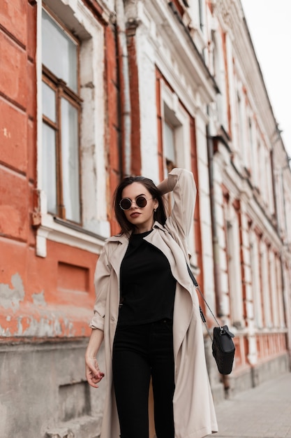 Giovane donna alla moda in occhiali da sole vintage in elegante trench in abiti casual neri con borsa in pelle nera cammina vicino al vecchio edificio in città. ragazza urbana di moda all'aperto. stile femminile giovanile.