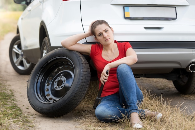 Treurige vrouw die op de grond zit bij een kapotte auto en op een reservewiel leunt