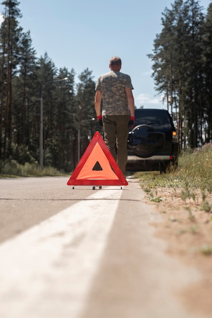 Segnale di attenzione triangolare sul lato della strada sulla linea bianca vicino all'auto rotta con l'autista che si allontana