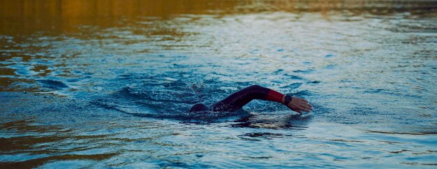 トライアスロン選手が日出時にウェットスーツを着て湖で泳いでいます