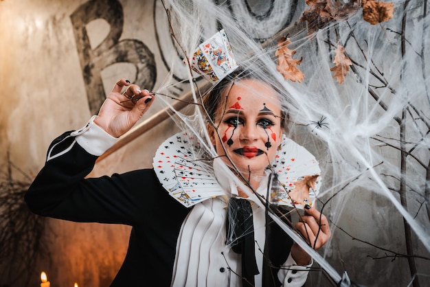 Trick or treat. Portrait of young woman with spooky makeup wearing Queen of Hearts costume with card collar looking at camera through spider web while visiting night Halloween party