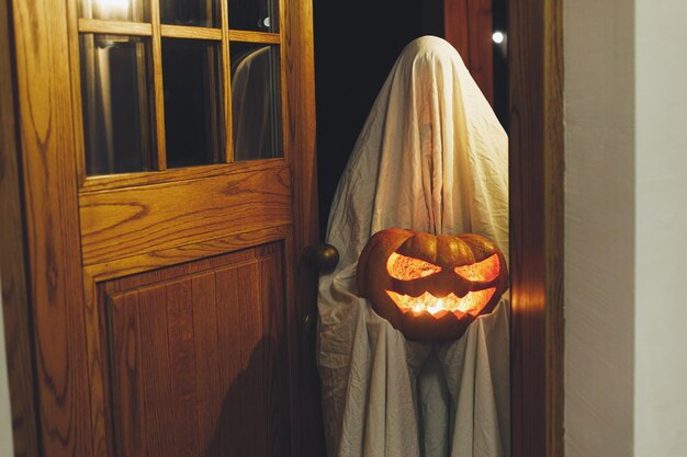 Foto dolcetto o scherzetto fantasma spettrale che tiene una zucca jack o lantern luminosa nelle porte al buio persona spaventosa e atmosferica di halloween vestita da fantasma dolcetto o scherzetto buon halloween