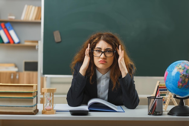 triest greep het hoofd van een jonge vrouwelijke leraar met een bril die aan het bureau zit met schoolhulpmiddelen in de klas